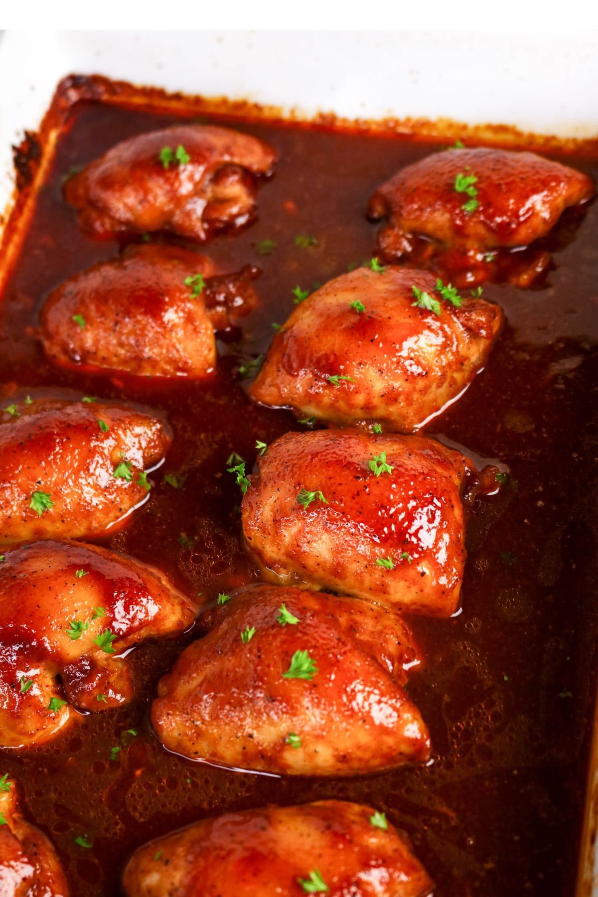 Baked chicken thighs coated in a glossy barbecue sauce, garnished with chopped parsley, arranged in a white baking dish.