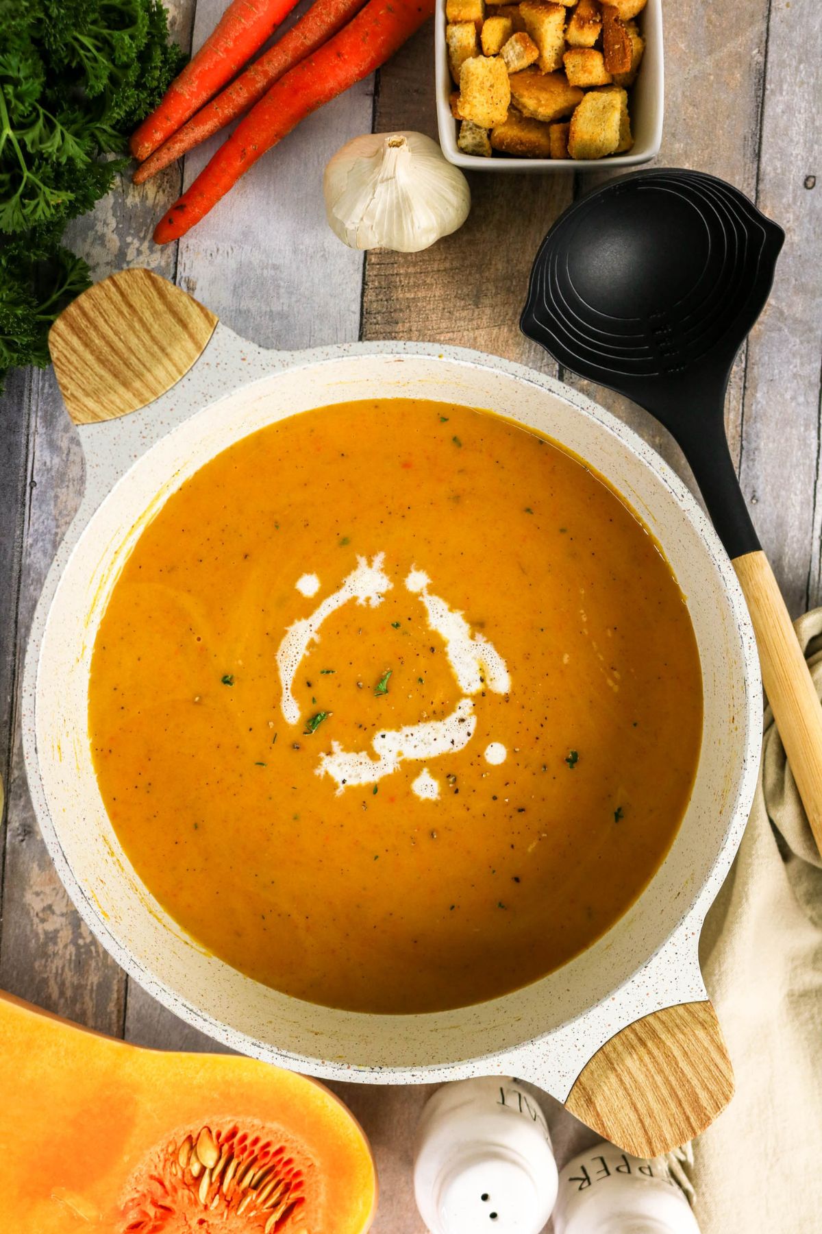A pot of creamy orange soup garnished with a swirl of cream, surrounded by carrots, garlic, croutons, fresh herbs, and a black ladle on a wooden table.