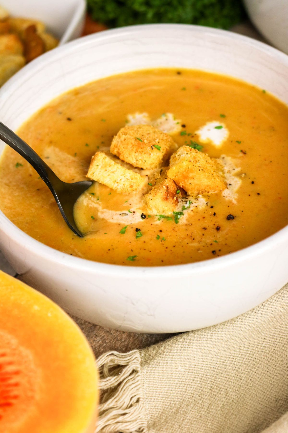 A bowl of creamy orange soup topped with croutons, black pepper, and herbs, with a spoon in the bowl and a beige napkin underneath.