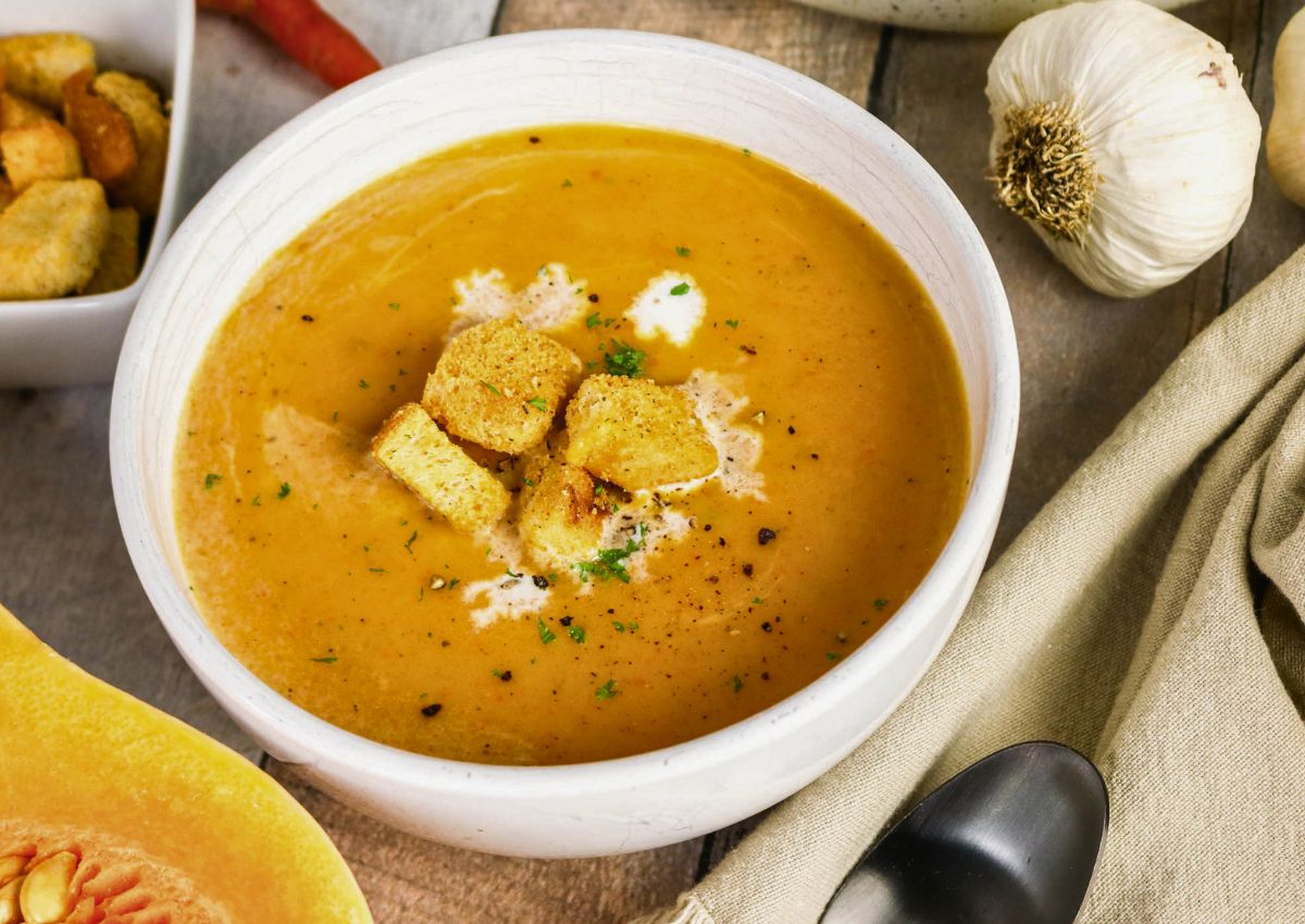 A bowl of pumpkin soup topped with croutons and cream, with garlic, a napkin, and a spoon nearby on a wooden table.