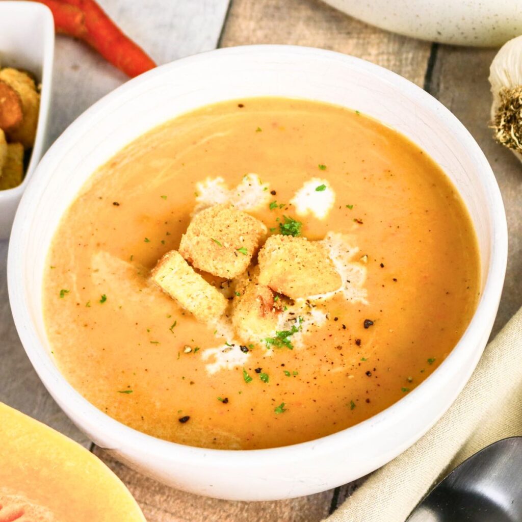 Bowl of creamy soup topped with croutons and herbs, with a spoon, garlic, and peppers visible nearby on a wooden surface.