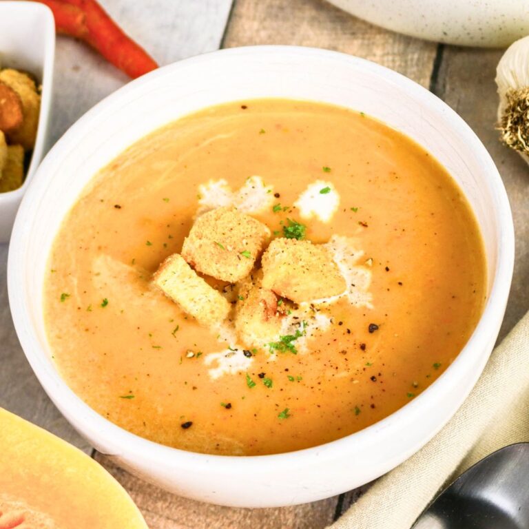 Bowl of creamy soup topped with croutons and herbs, with a spoon, garlic, and peppers visible nearby on a wooden surface.