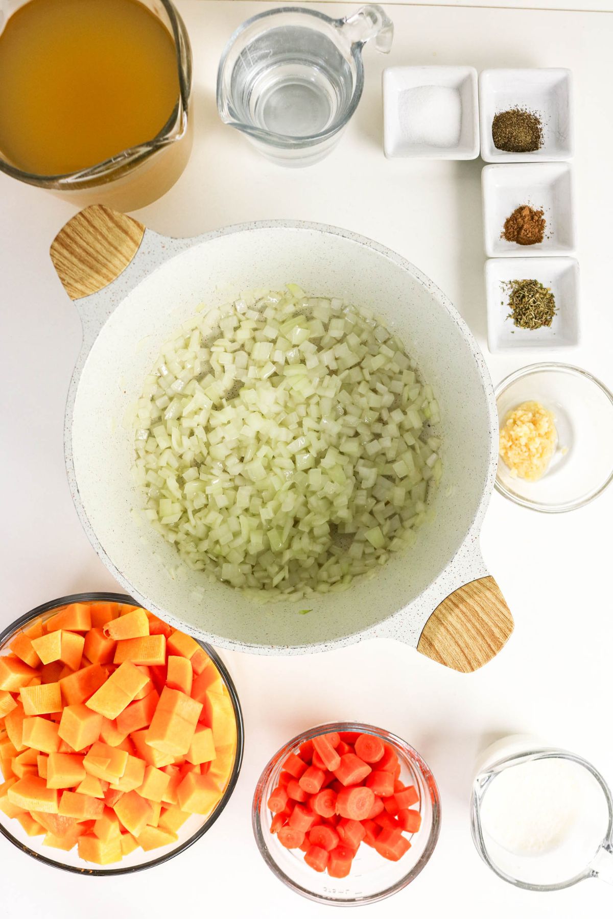 Chopped onions in a pot, with bowls of diced sweet potatoes, carrots, garlic, broth, water, spices, salt, and pepper arranged on a white surface.