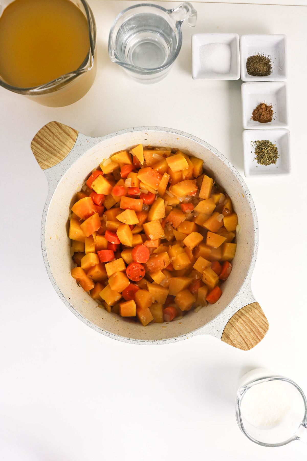 A pot of chopped carrots and root vegetables sits on a white surface, surrounded by glass containers of broth, water, seasonings, and three small dishes with salt, pepper, and herbs.