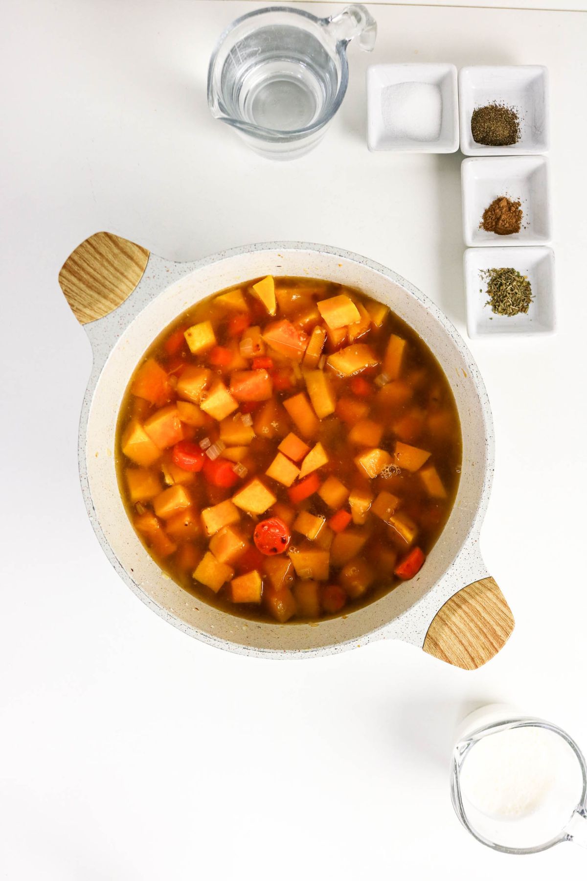 A pot of vegetable soup sits on a white surface next to a cup of water, spices in small bowls, and a glass of salt.