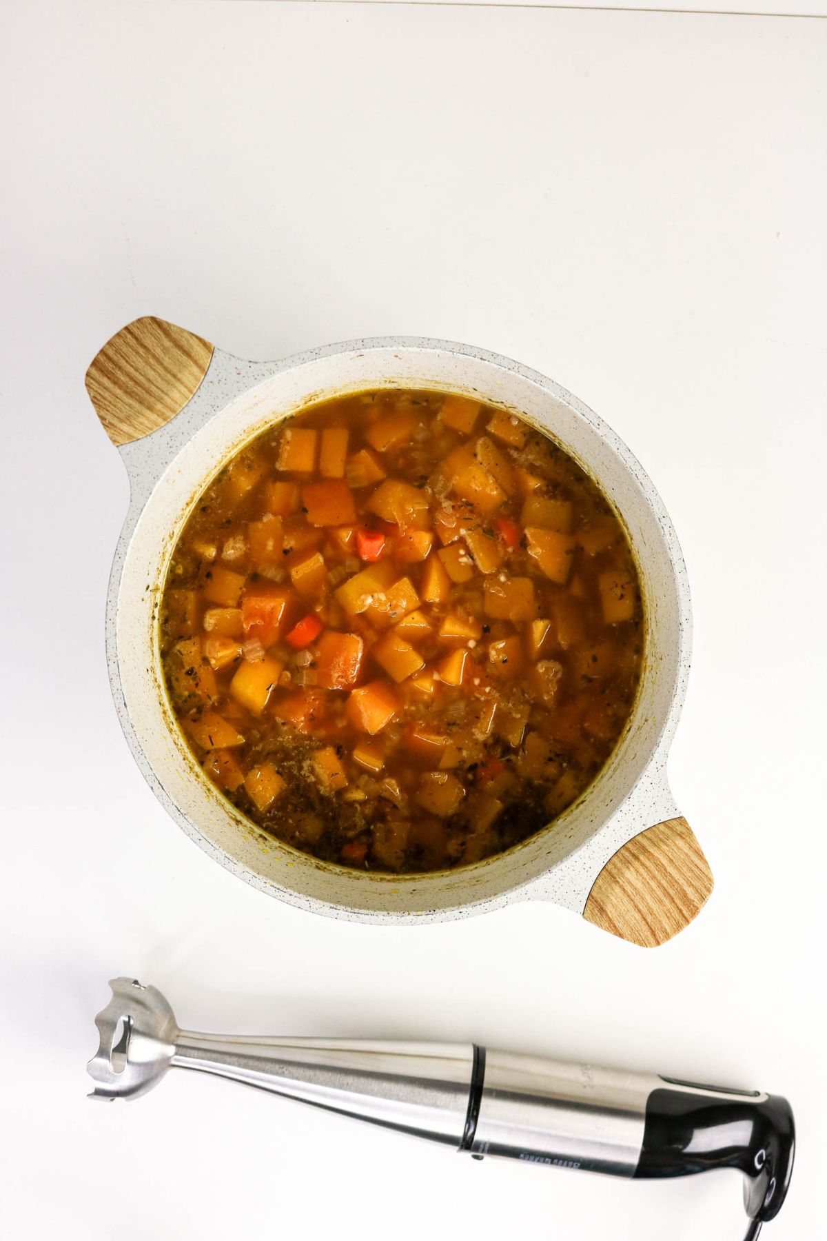 A pot of diced vegetables in broth sits on a white surface next to an immersion blender.