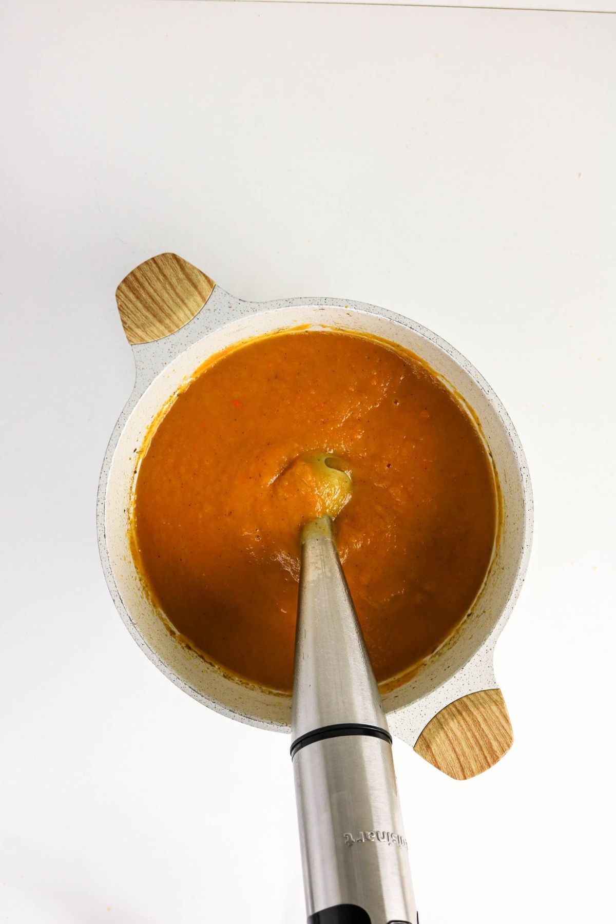A pot of orange soup is being blended with an immersion blender on a white surface.
