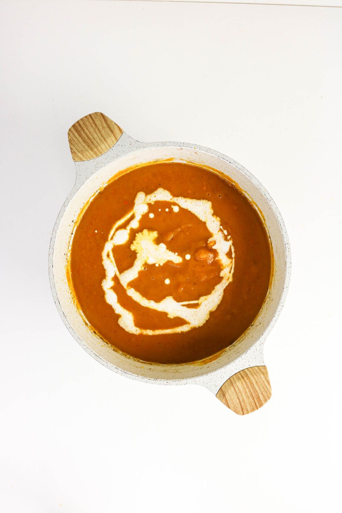 White pot with wooden handles containing orange soup topped with a swirl of cream, placed on a white background.