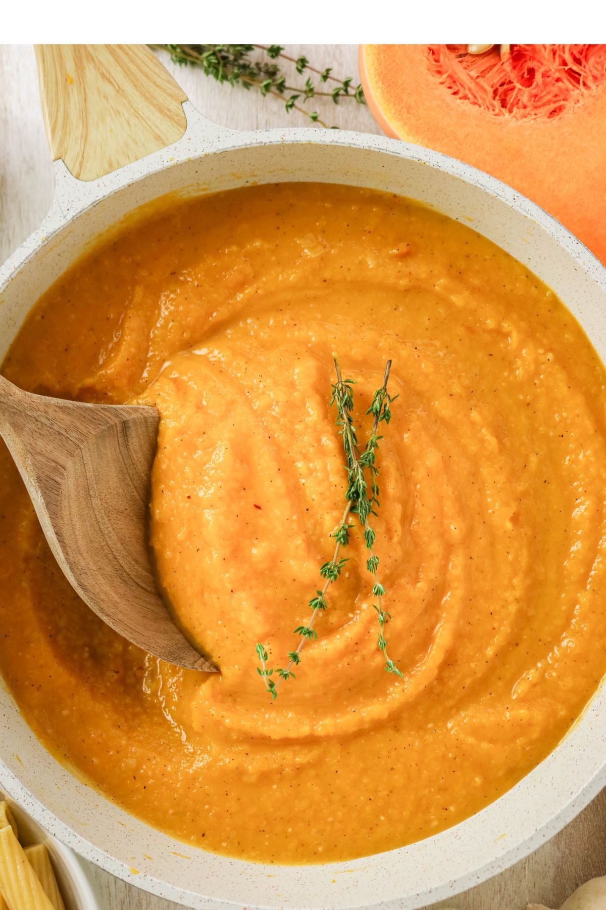 A white saucepan filled with creamy orange soup, garnished with fresh thyme sprigs, and stirred with a wooden spoon.