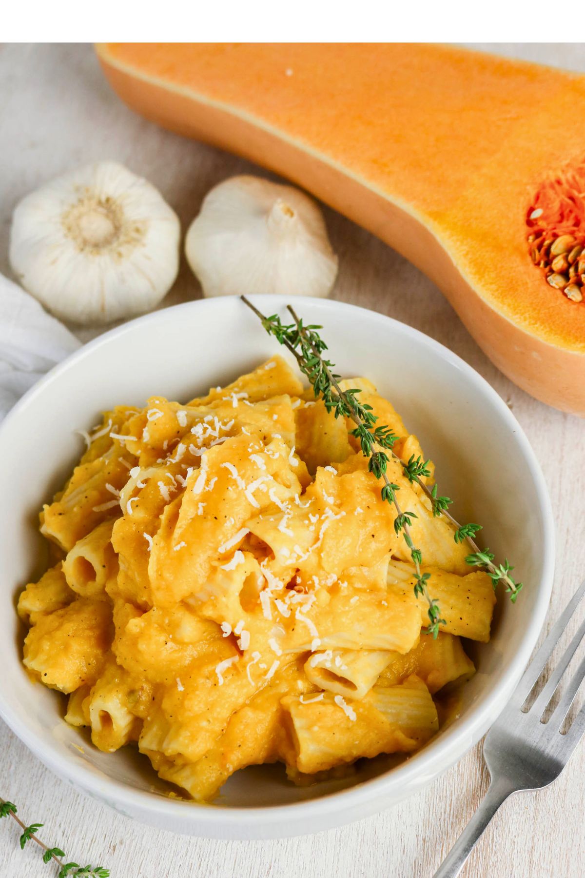 Bowl of rigatoni pasta with creamy orange sauce, garnished with grated cheese and thyme; garlic bulbs and a halved butternut squash in the background.