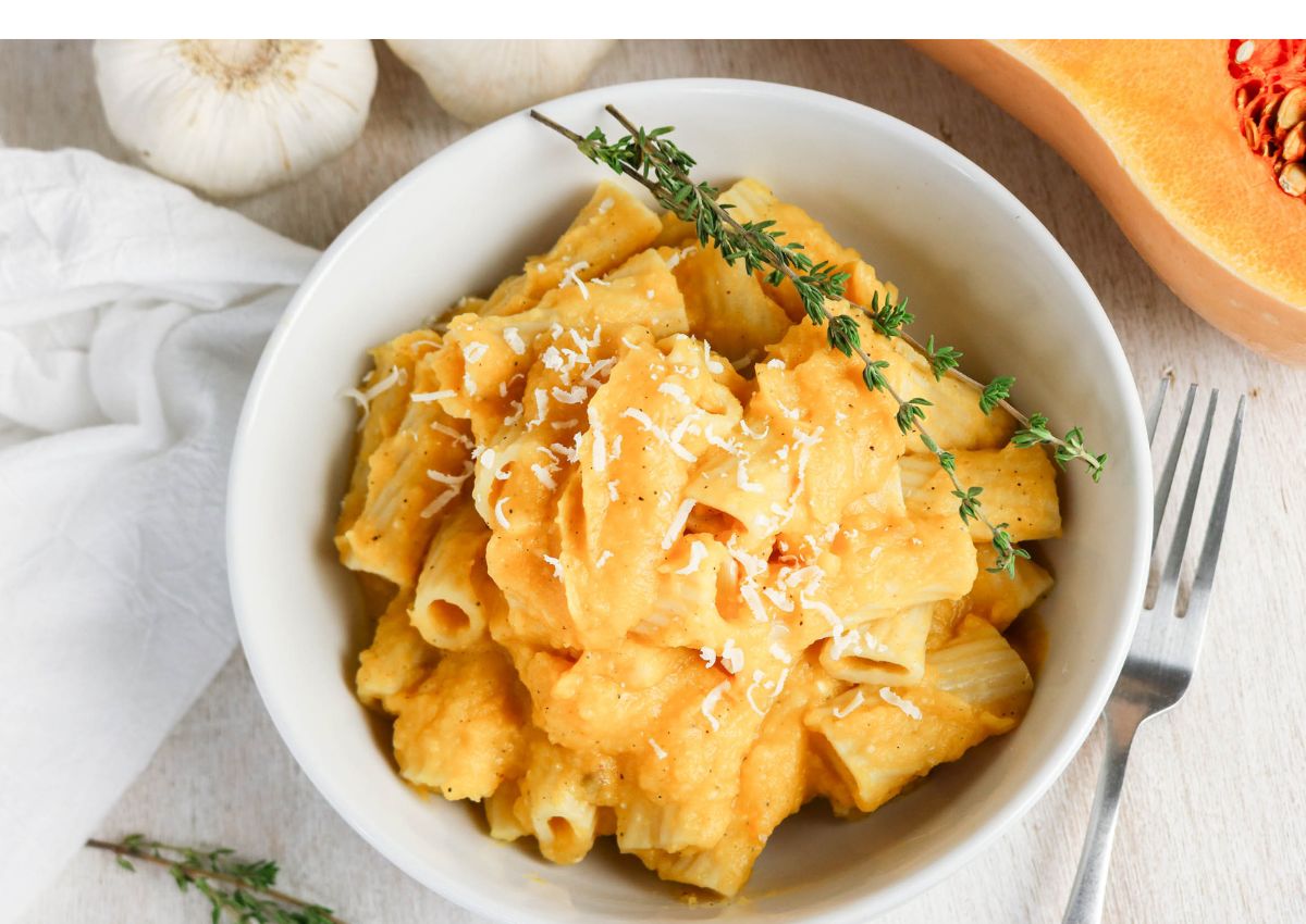 Bowl of rigatoni pasta in a creamy orange sauce, topped with grated cheese and a sprig of thyme, with a fork and garlic visible nearby.