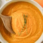 Creamy orange soup in a pot, stirred with a wooden spoon, garnished with fresh thyme sprigs.