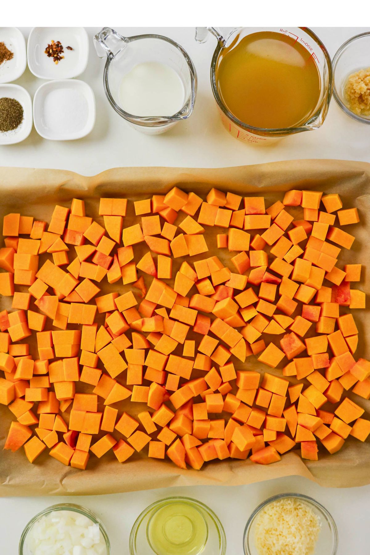 Diced butternut squash on a parchment-lined baking sheet, surrounded by bowls of ingredients including broth, milk, oil, onions, garlic, spices, and shredded cheese.