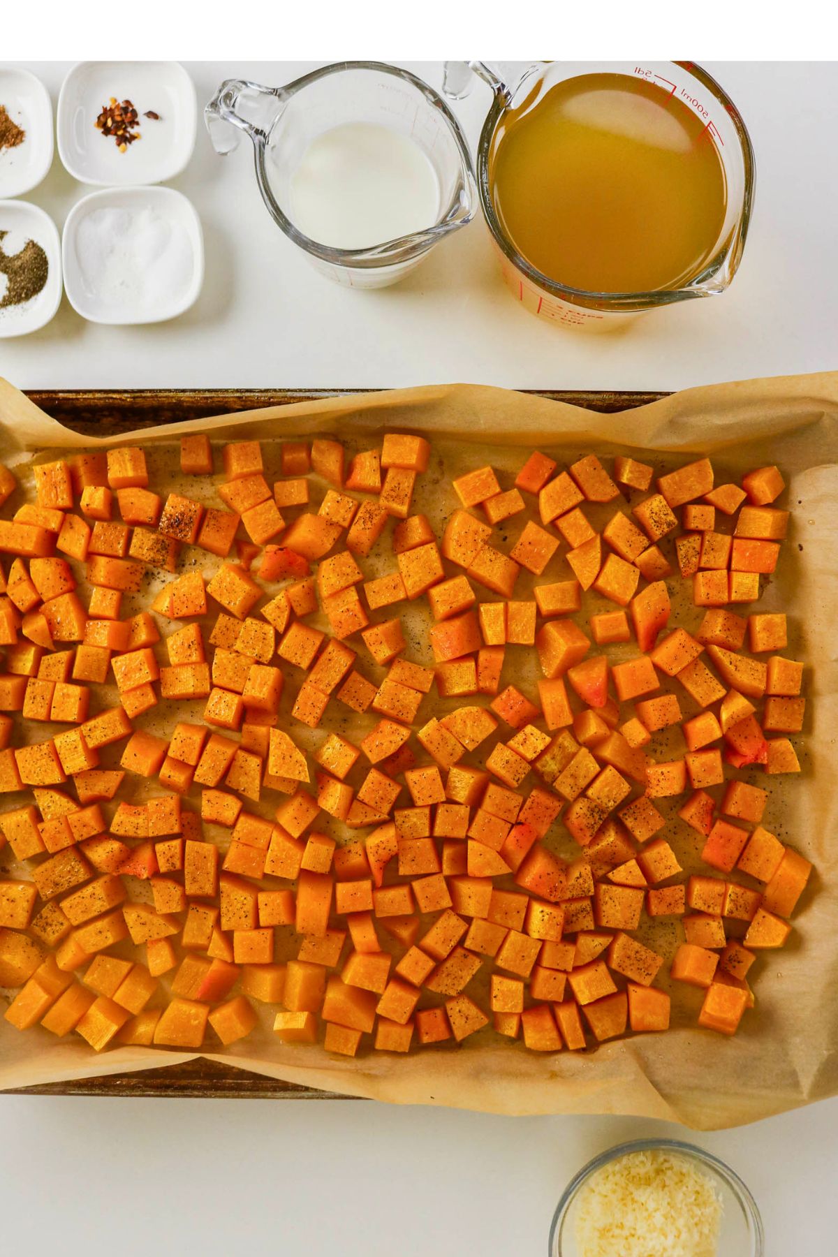 Cubed sweet potatoes on a parchment-lined baking sheet, surrounded by bowls of spices, shredded cheese, cream, and a measuring cup of broth on a white surface.