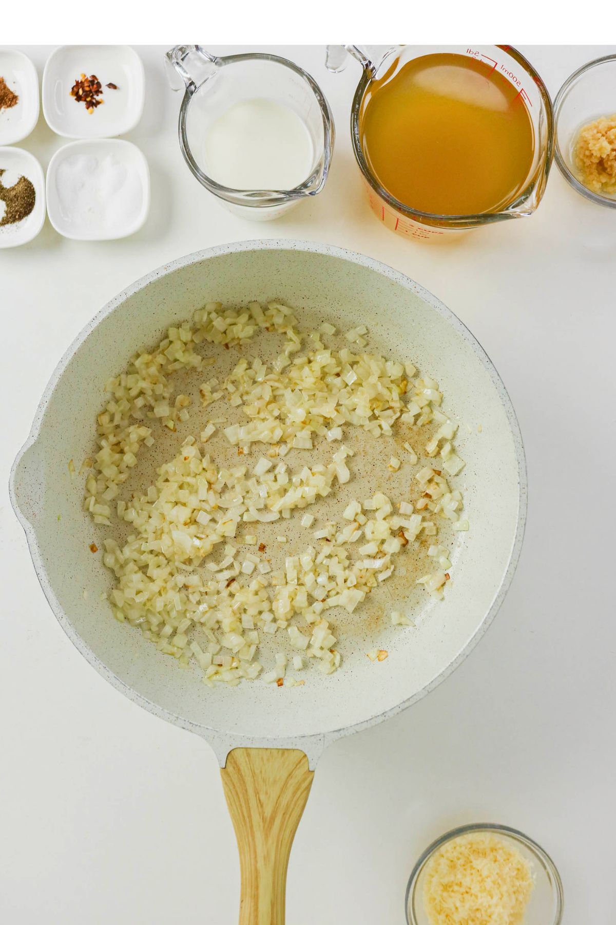 A white pan with sautéed onions, surrounded by bowls of spices, grated cheese, milk, and measuring cup of broth on a white surface.