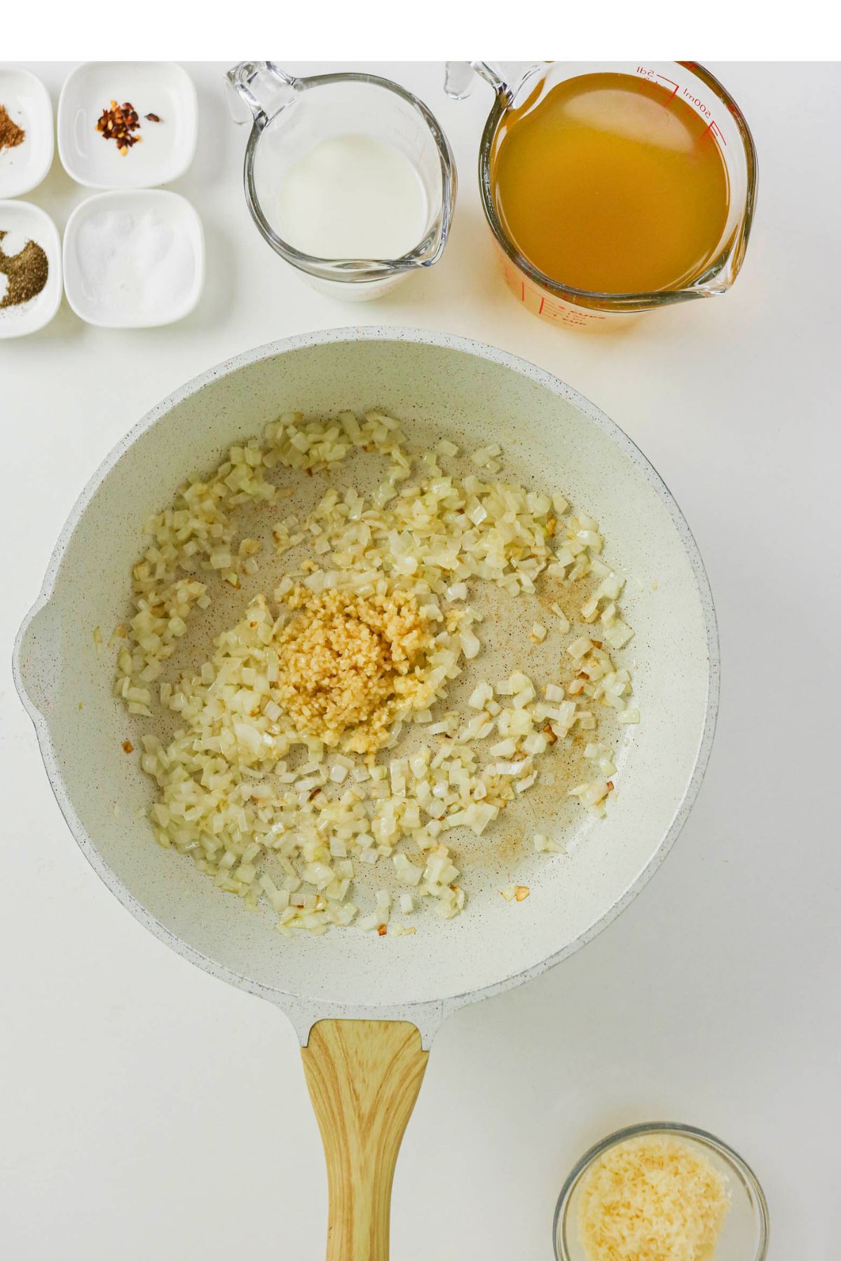 A frying pan with chopped onions and minced garlic, surrounded by bowls of spices, grated cheese, milk, and a measuring cup of broth on a white background.