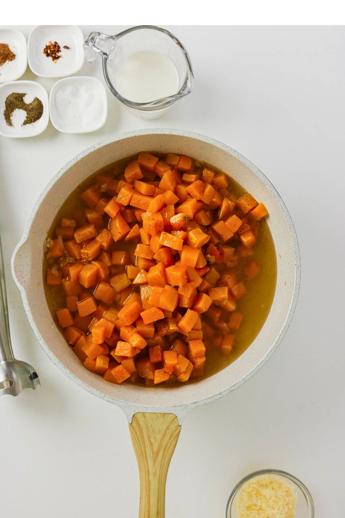 A white pan with diced sweet potatoes in broth sits on a white surface, surrounded by small bowls of spices, a glass of milk, and shredded cheese.