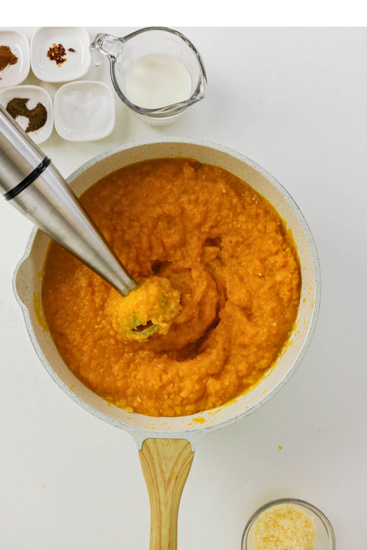 An immersion blender blends orange soup in a white pot, surrounded by a cup of milk, seasonings in small bowls, and a glass bowl with shredded cheese on a white surface.