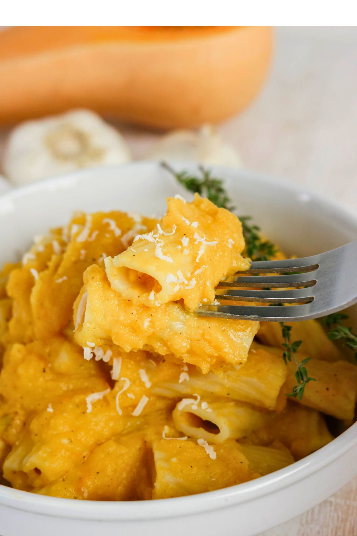 A fork holds rigatoni pasta covered in a creamy orange sauce, topped with shredded cheese and garnished with fresh herbs in a white bowl.