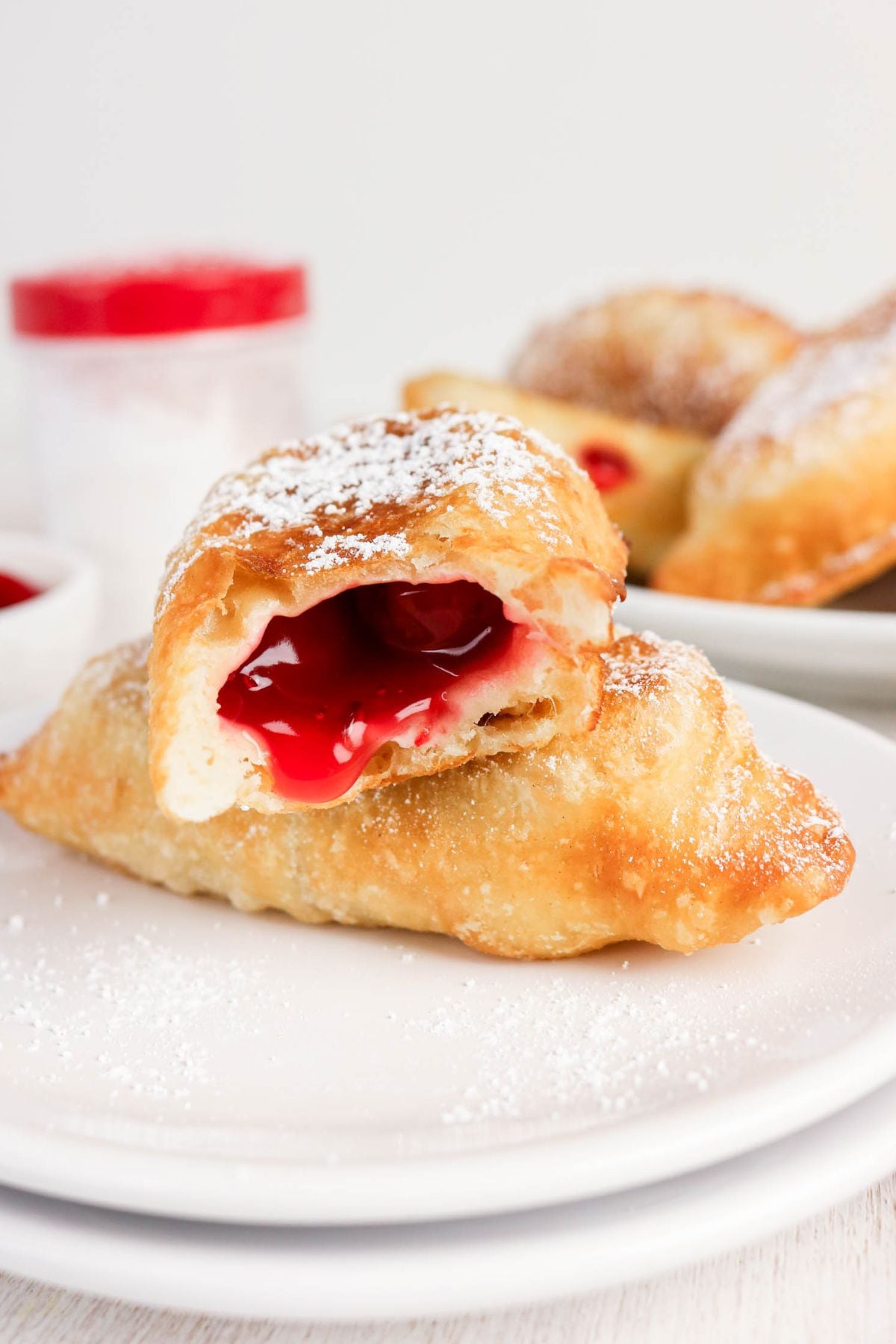 A golden, flaky pastry filled with bright red cherry filling, dusted with powdered sugar, sits on a white plate with another whole pastry and a jar in the background.