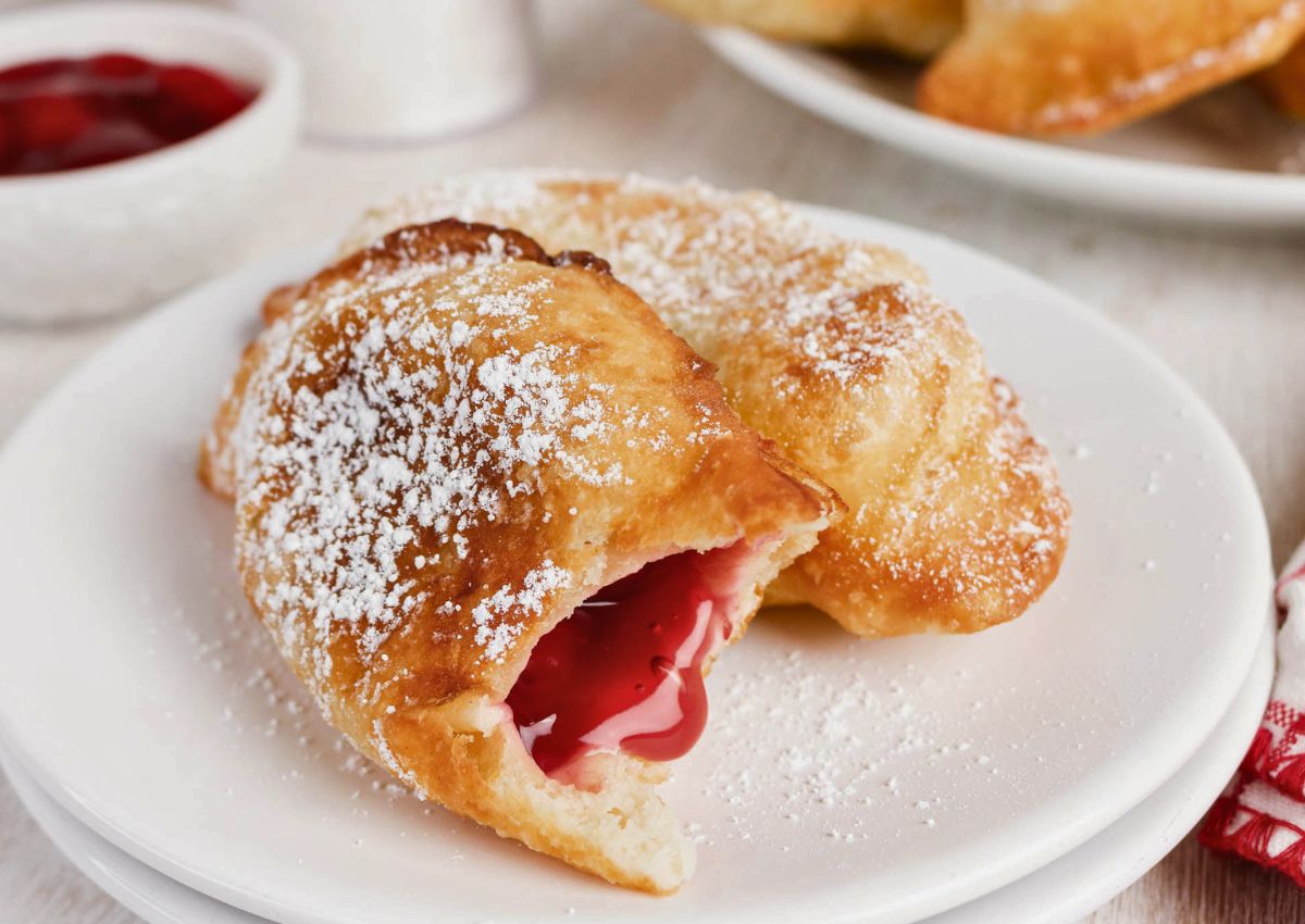 Two cherry-filled fried pies on a white plate, dusted with powdered sugar. One pie is partially broken open, showing the red cherry filling inside.