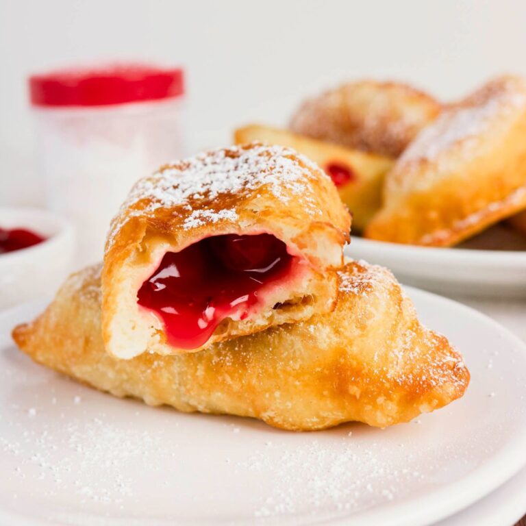 Cherry-filled fried pastry topped with powdered sugar, one pastry cut open to show the bright red filling, served on a white plate.