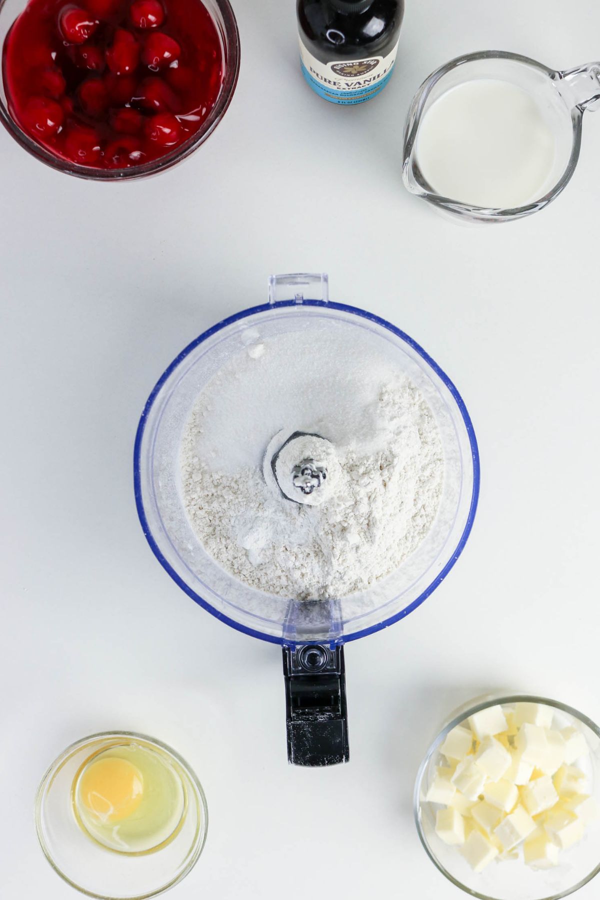 Overhead view of a food processor with flour and sugar, surrounded by bowls of cubed butter, an egg, cherry filling, vanilla extract, and milk on a white surface.