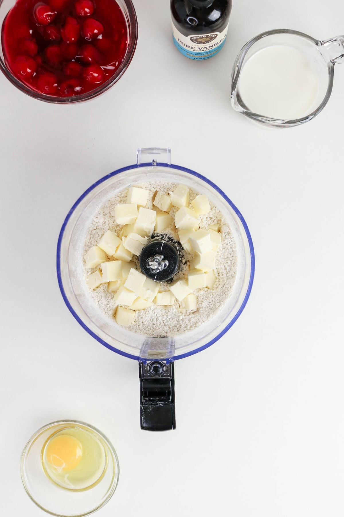 Overhead view of a food processor with flour and cubed butter, surrounded by a bowl of cherries, a measuring cup of milk, a bottle of vanilla, and a bowl with a cracked egg.