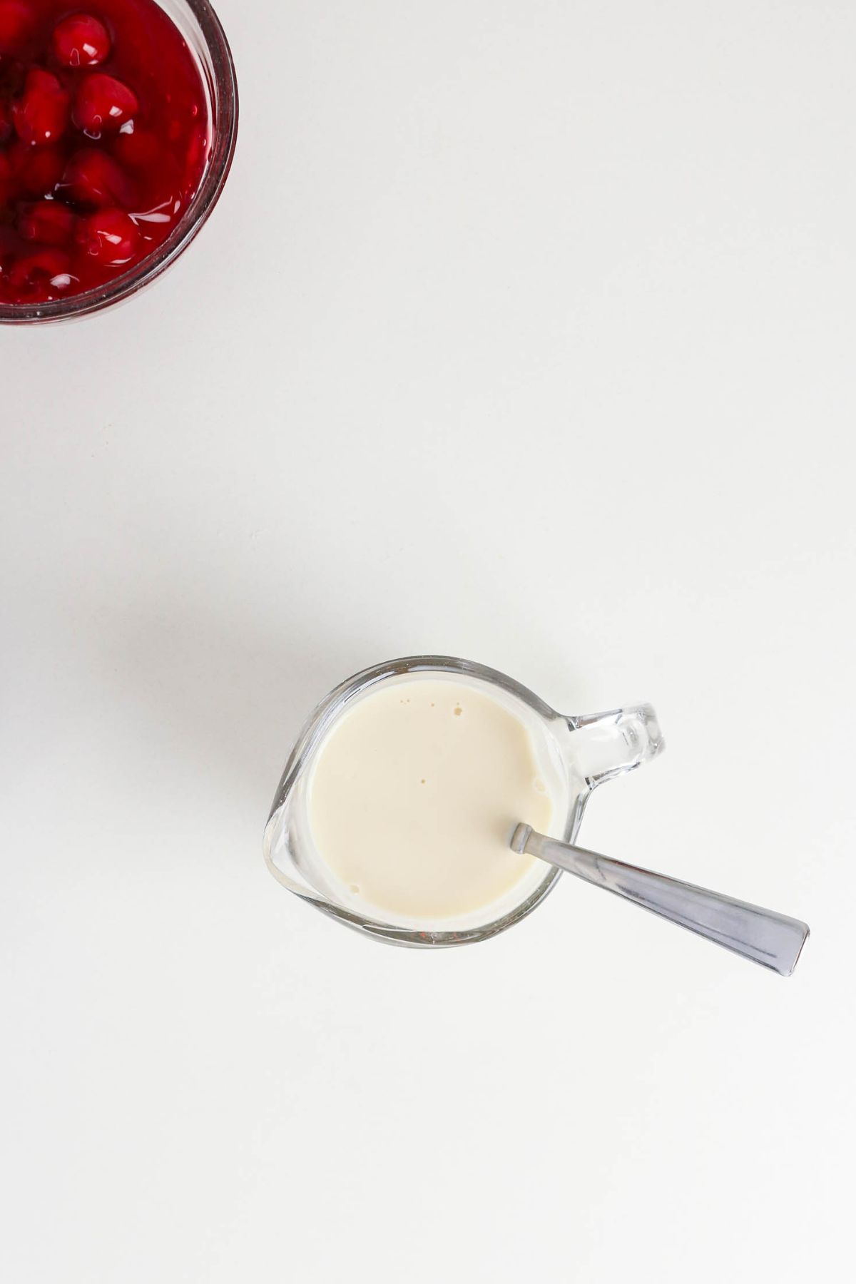 A glass measuring cup with cream and a spoon next to a partial bowl of cherry sauce on a white surface.