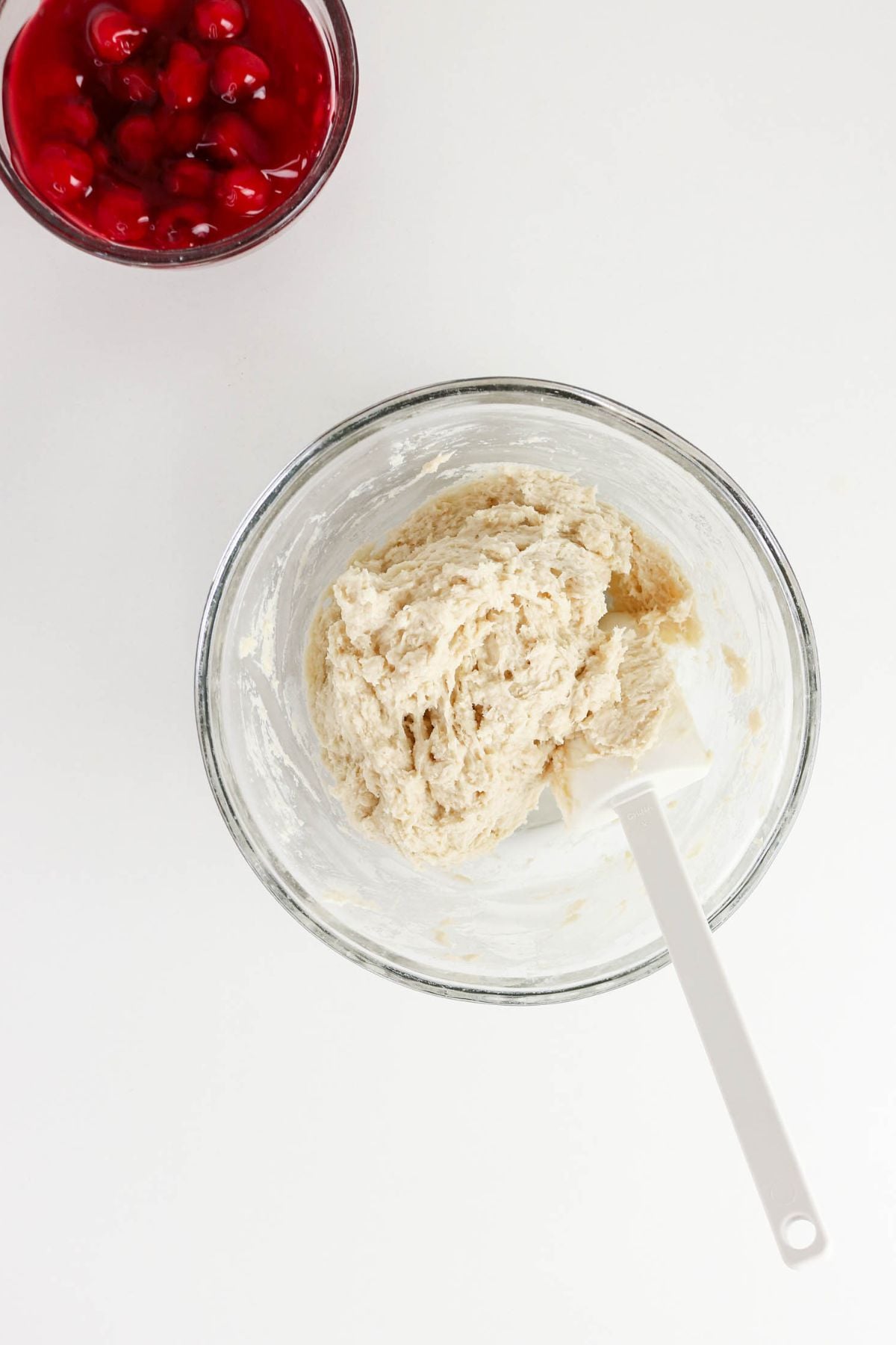 A glass bowl of raw dough with a white spatula next to a bowl of cherry topping on a white surface.