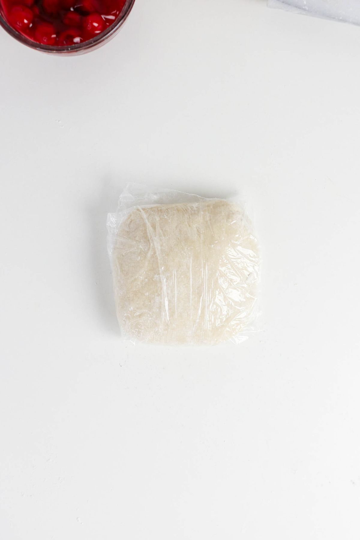 A square portion of dough wrapped in plastic wrap sits on a white surface. A bowl with red cherries is partially visible in the top left corner.