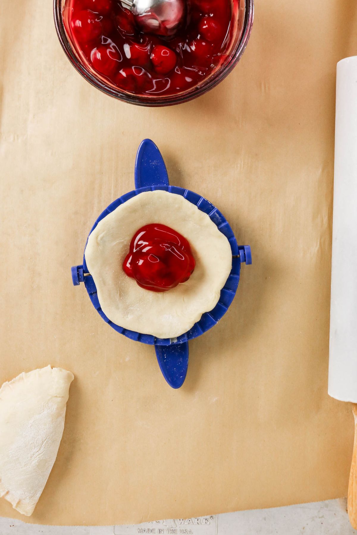 A blue pastry press holds dough with cherry filling inside, next to a bowl of cherry filling and a folded, uncooked hand pie on a parchment-lined surface.