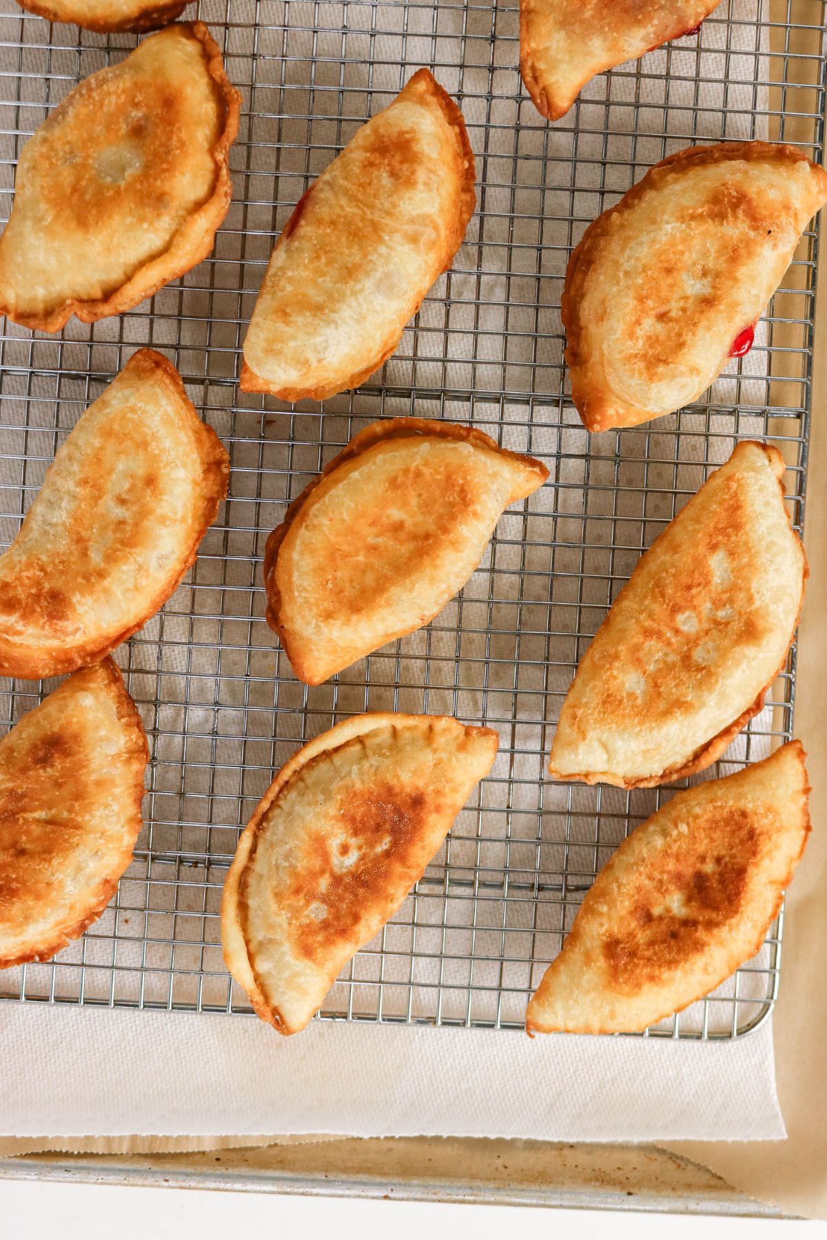 Golden brown, crescent-shaped empanadas cool on a wire rack placed over a paper towel-lined baking sheet.