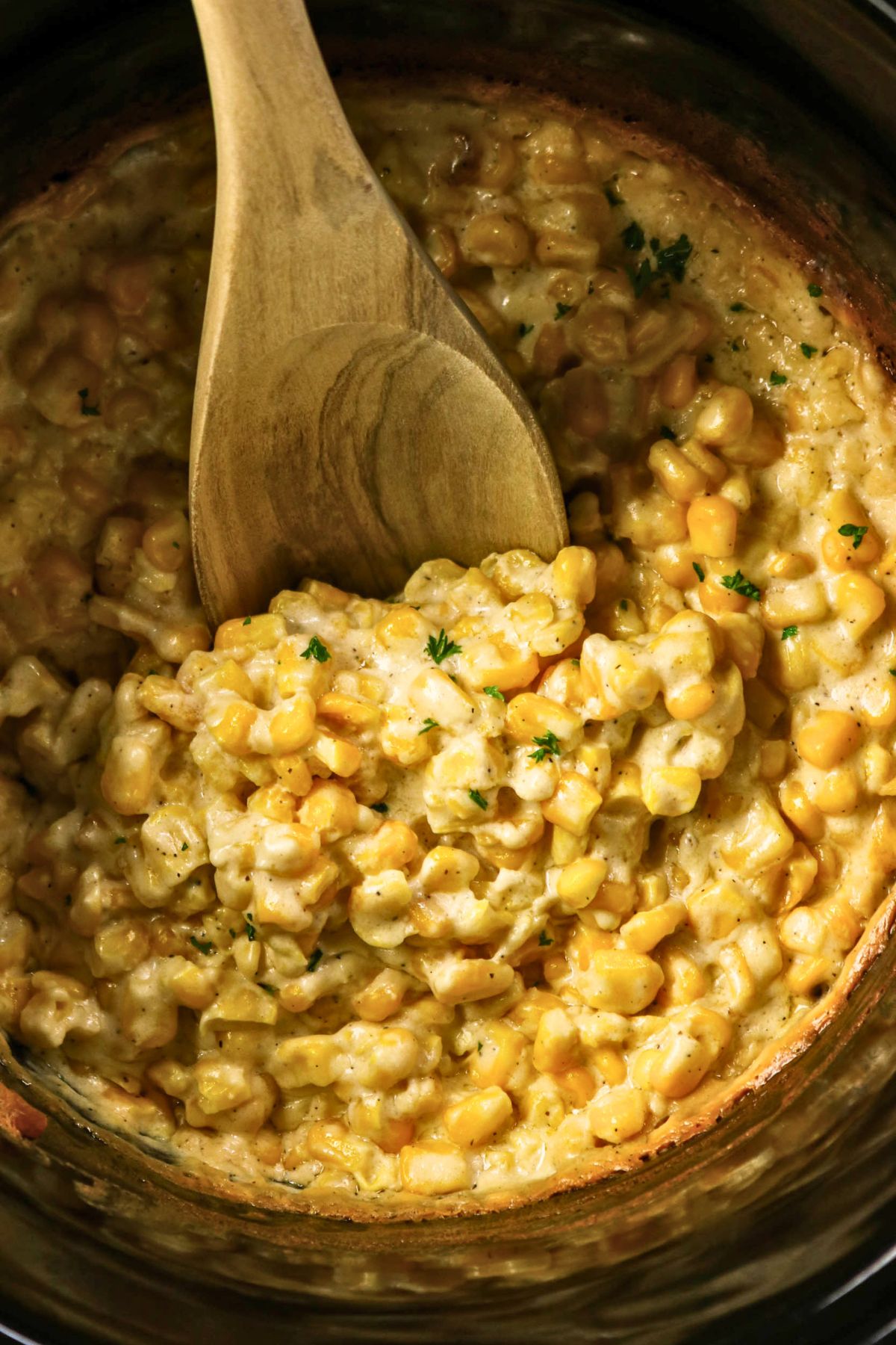 Creamy corn casserole with herbs, shown in a pot with a wooden spoon scooping the mixture.