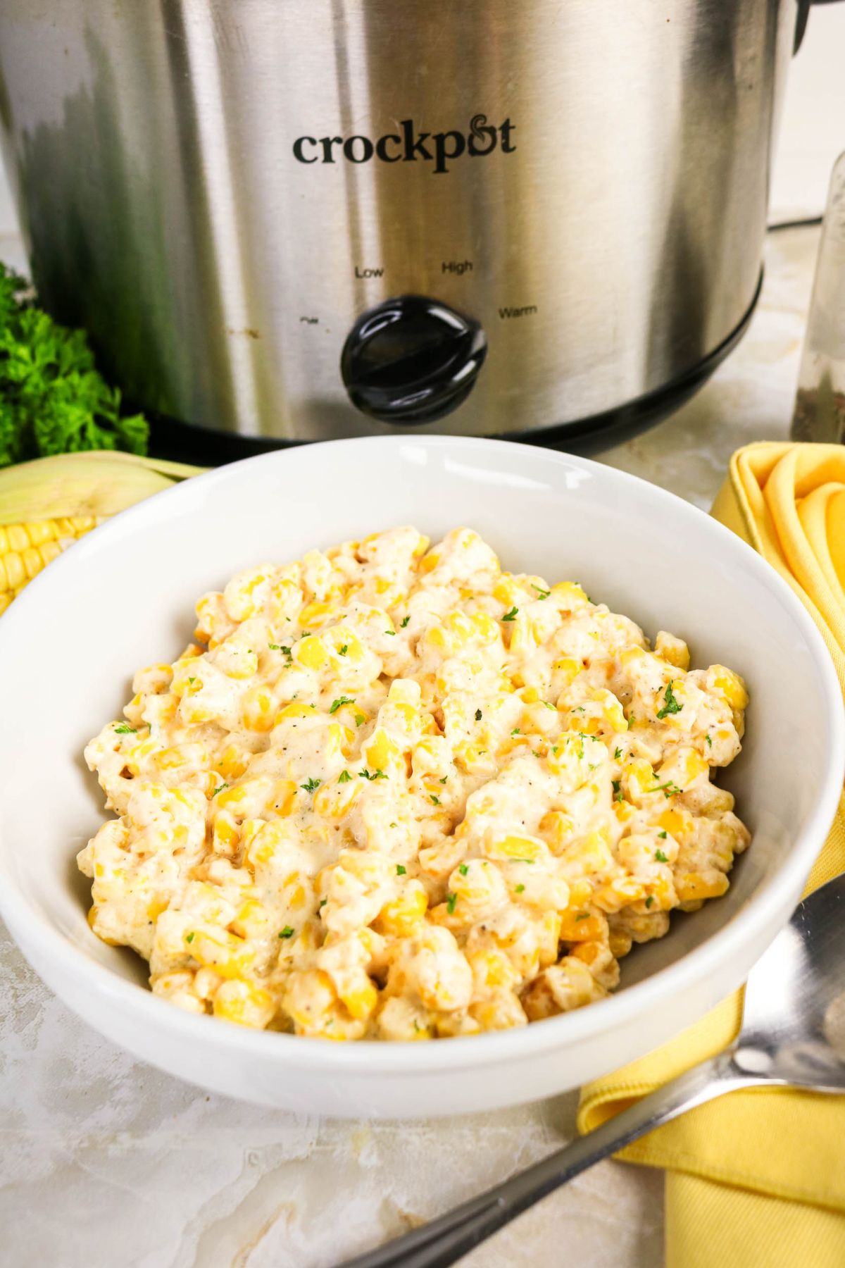 A white bowl of creamy corn sits in front of a slow cooker on a marble countertop, with a yellow napkin, spoon, corn, and parsley beside it.