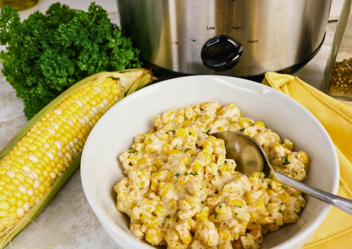 Bowl of creamy corn casserole with a spoon, next to fresh parsley, corn on the cob, and a slow cooker.
