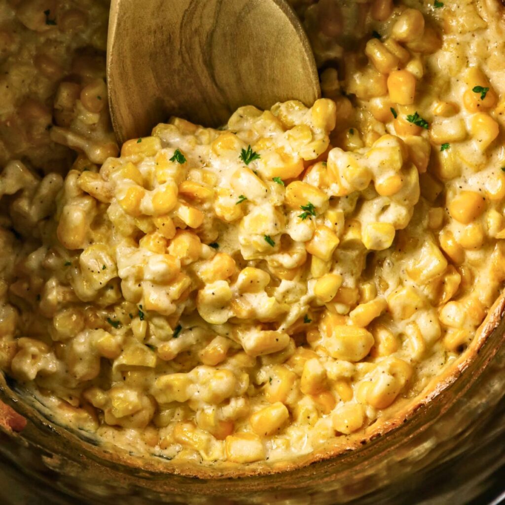 Creamed corn with visible corn kernels and creamy sauce, garnished with small bits of green herbs, being stirred with a wooden spoon.
