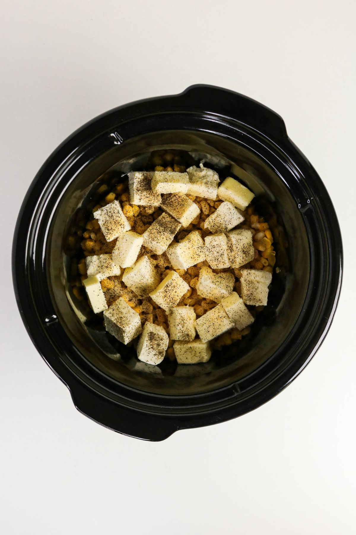 Overhead view of a slow cooker filled with corn, cubes of cream cheese and butter, topped with ground black pepper.