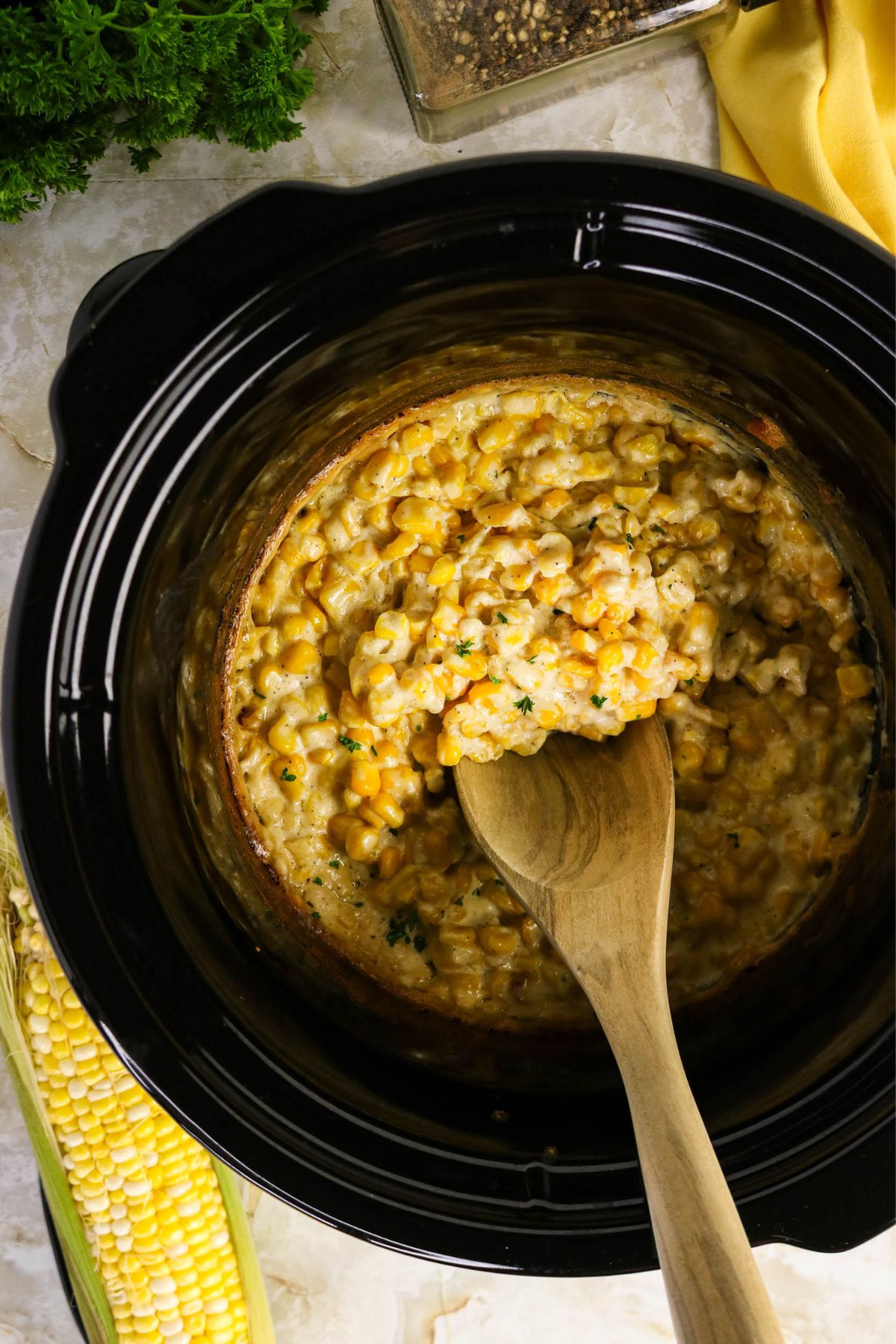 A wooden spoon rests in a slow cooker filled with creamy corn, surrounded by fresh corn, parsley, and a yellow cloth.