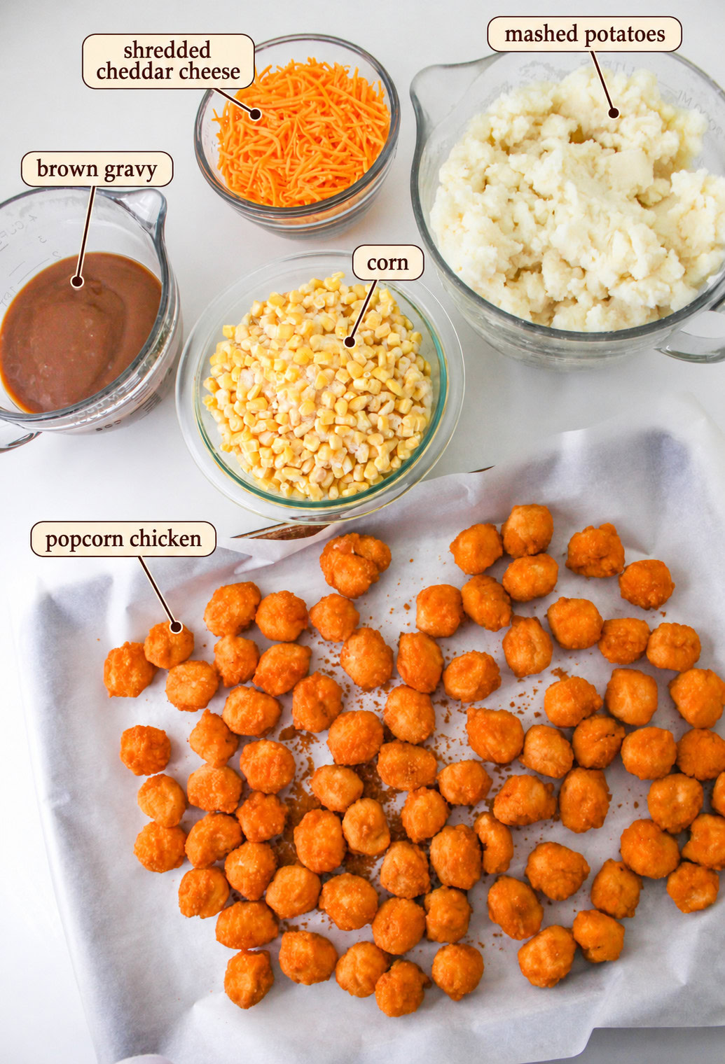 Ingredients for a KFC bowl casserole: bowls of shredded cheddar, mashed potatoes, brown gravy, and corn next to a baking sheet of cooked popcorn chicken.