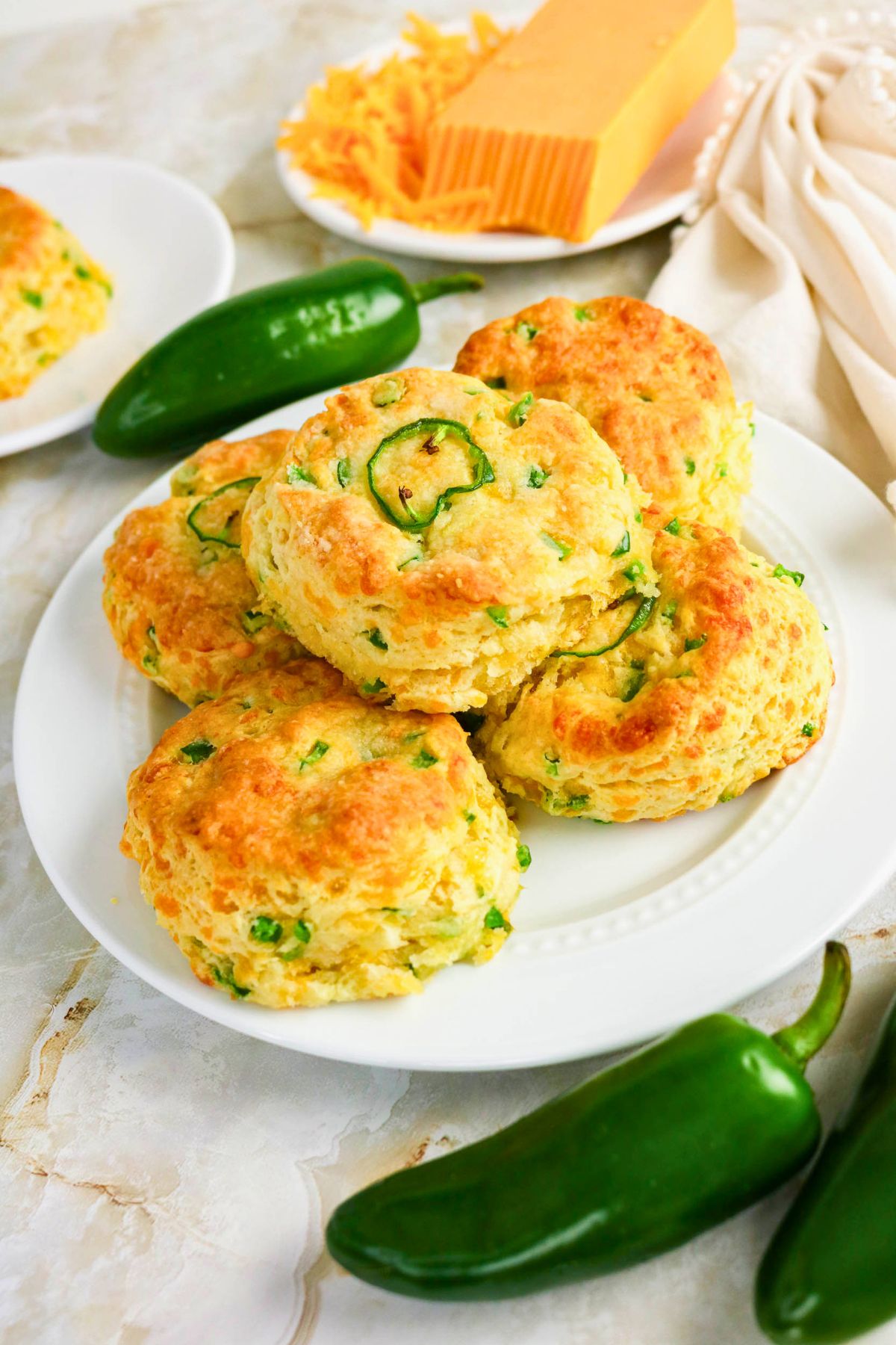 Plate of golden biscuits with chopped green jalapeños, surrounded by whole jalapeños, shredded cheese, and a block of cheddar on a marble surface.