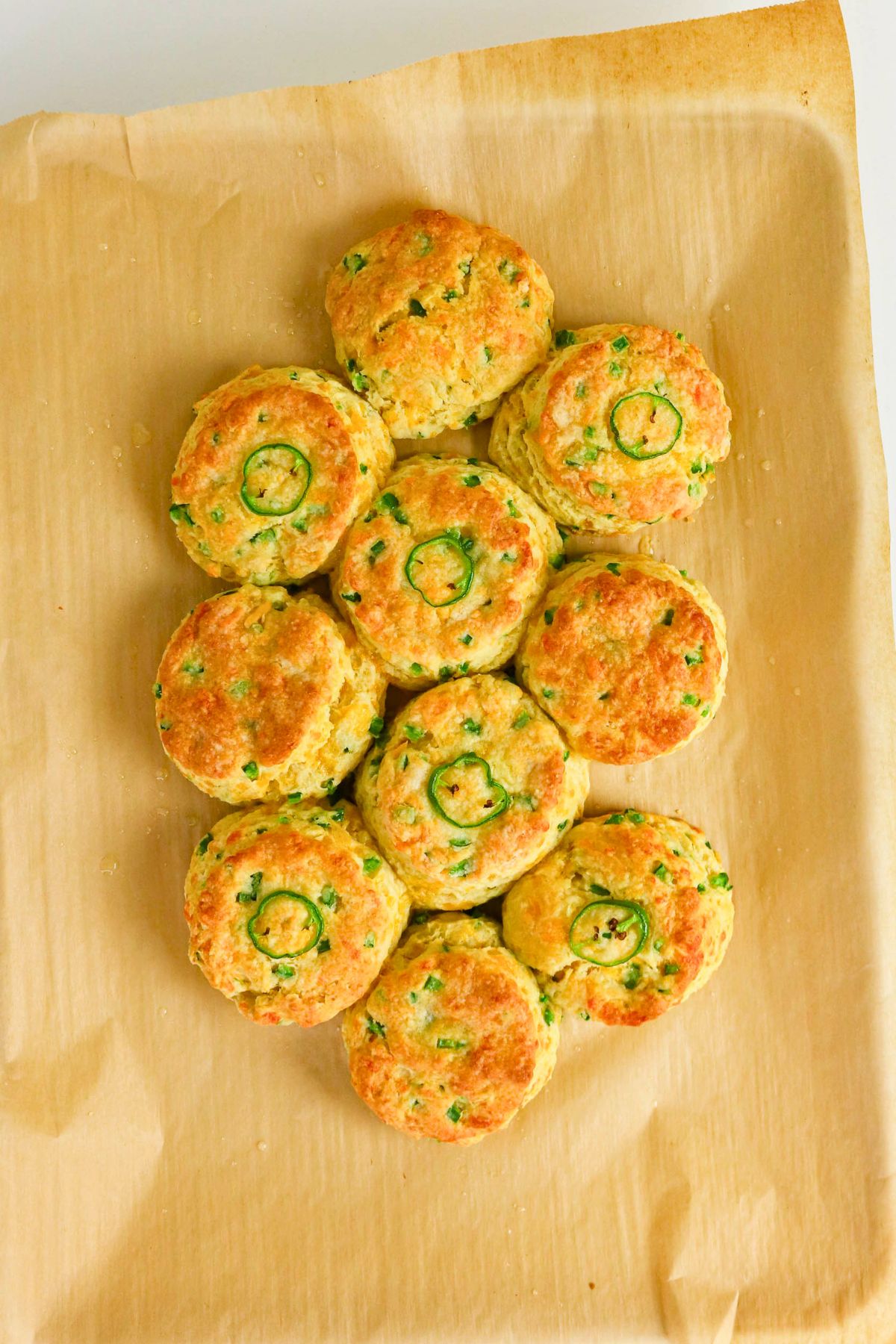 Nine baked biscuits with chopped green herbs and jalapeno slices on top, arranged on parchment paper.