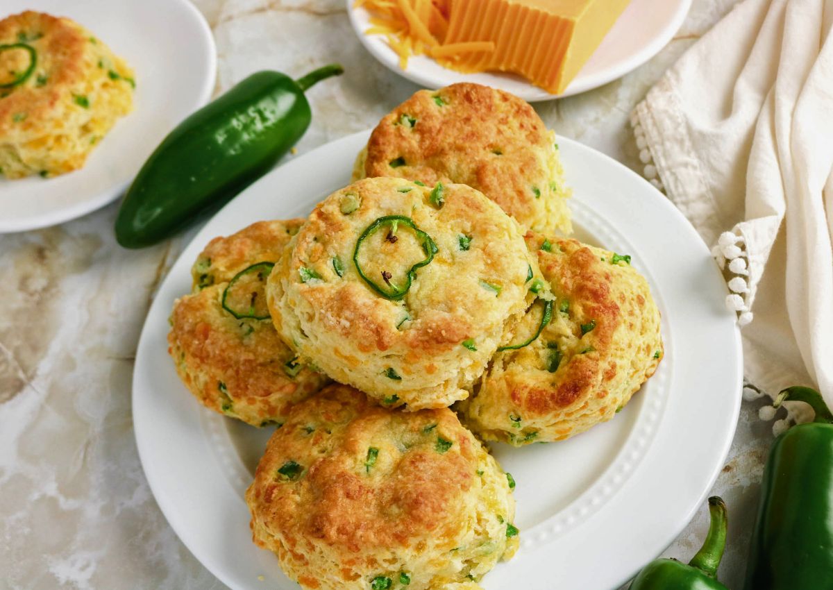 A plate of golden-brown biscuits with jalapeño slices on top, surrounded by whole jalapeños and a block of cheddar cheese on a marble surface.