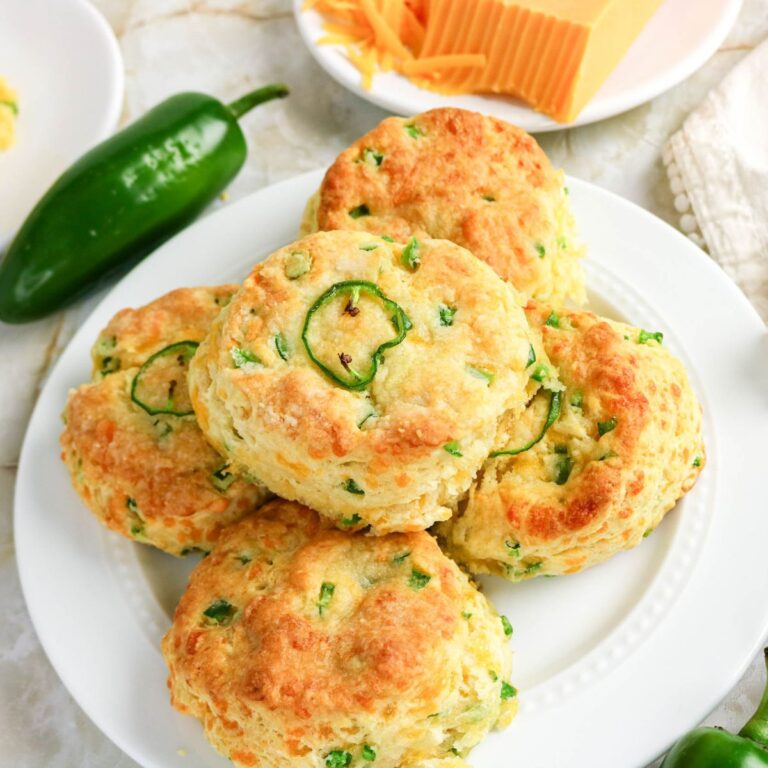 Five jalapeño cheddar biscuits are stacked on a white plate, surrounded by a block of cheddar cheese, sliced cheese, a jalapeño pepper, and a white napkin.