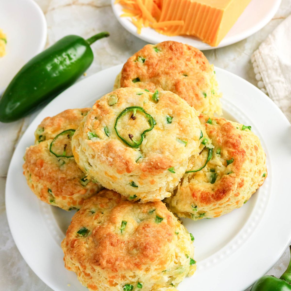 Five jalapeño cheddar biscuits are stacked on a white plate, surrounded by a block of cheddar cheese, sliced cheese, a jalapeño pepper, and a white napkin.