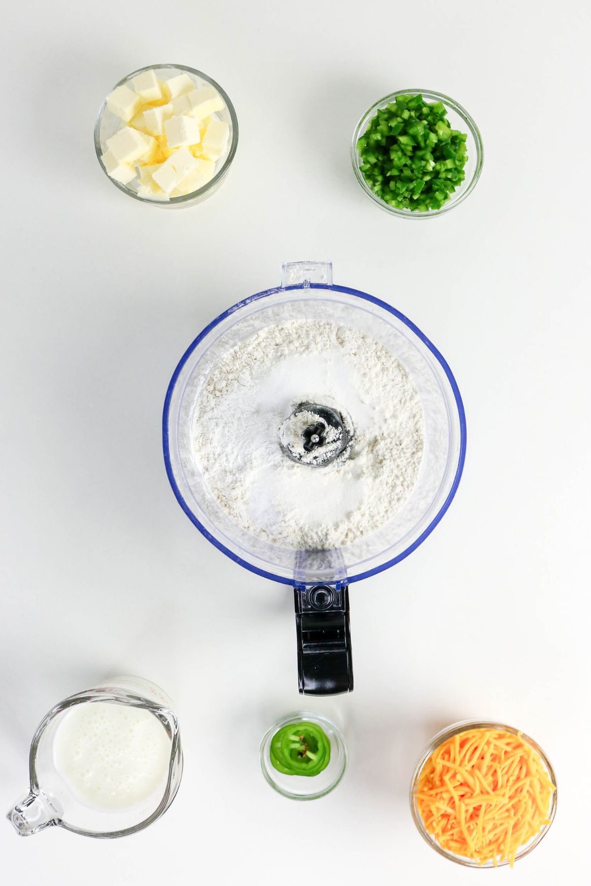 Top view of a food processor with flour, surrounded by bowls of cubed butter, chopped jalapeños, shredded cheese, a sliced jalapeño, and a measuring cup of buttermilk on a white background.