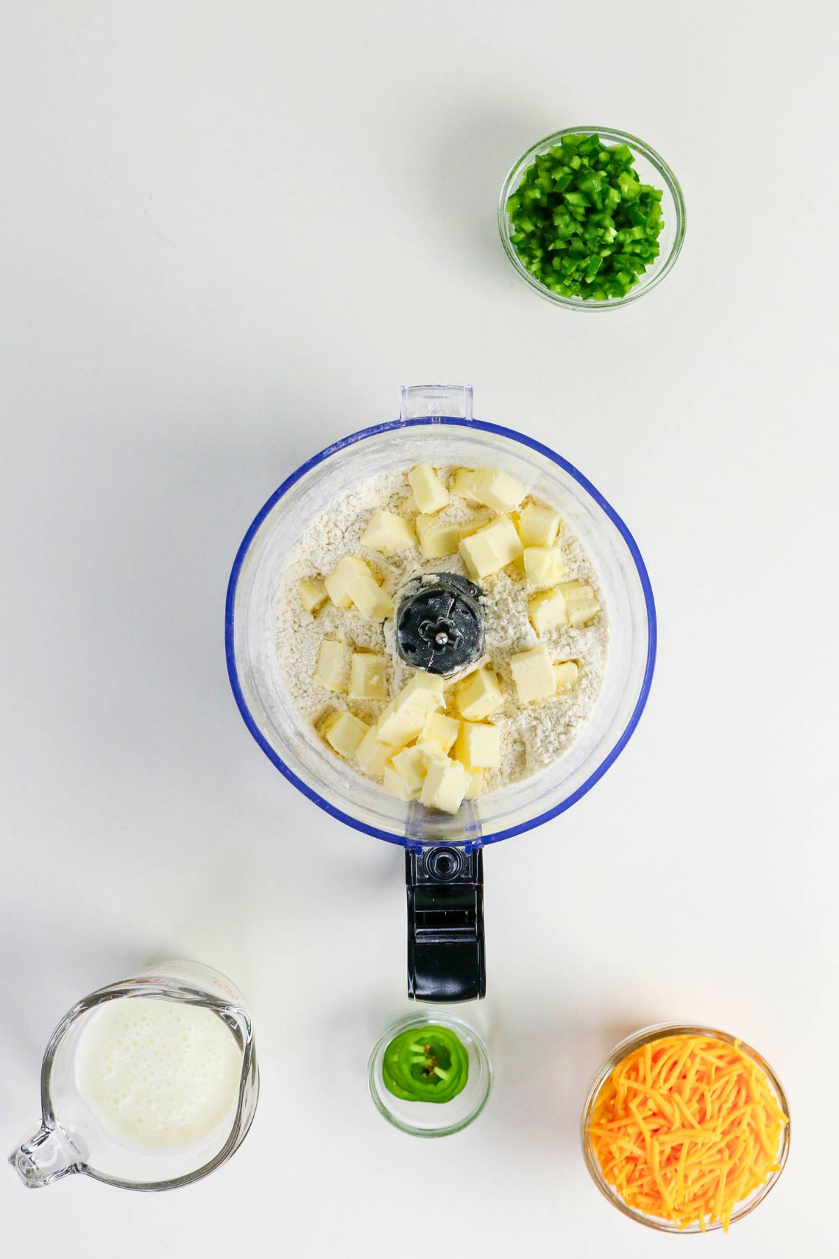 A food processor with flour and butter cubes, surrounded by bowls of chopped jalapeño, shredded cheese, sliced jalapeño, and a measuring cup of buttermilk on a white surface.