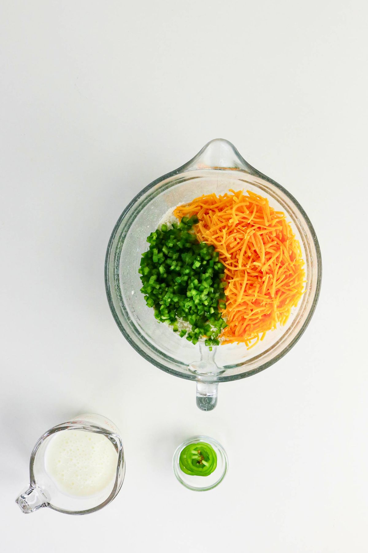 Glass bowl with shredded cheddar cheese and chopped green peppers, next to a small pitcher of milk and a jar with sliced jalapeños on a white background.