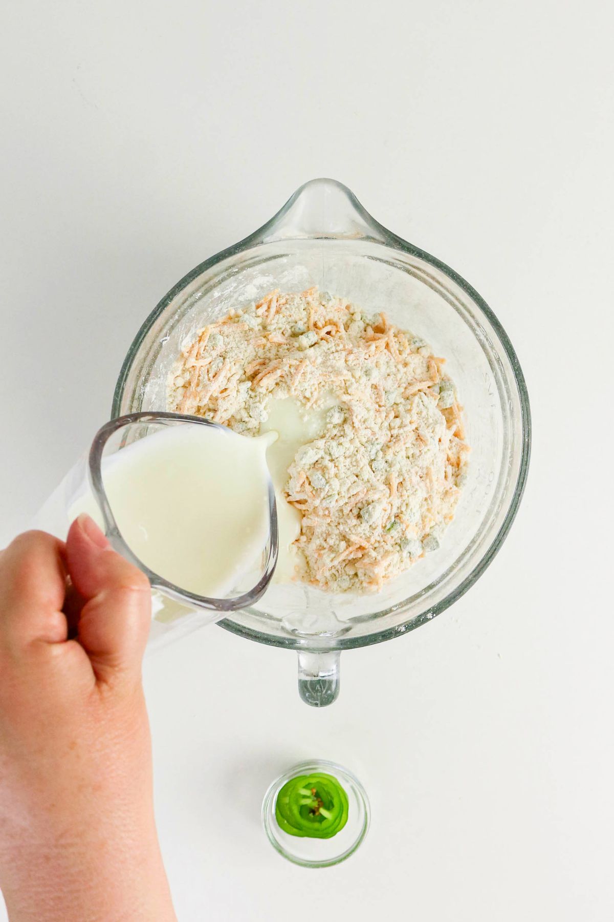 A hand pours milk from a measuring cup into a glass mixing bowl containing flour and shredded cheese. A small bowl with a green ingredient sits nearby.