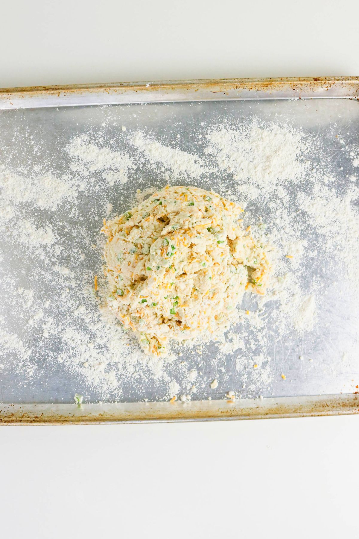 A mound of flour-dusted dough with visible green and orange bits rests on a floured metal baking sheet.